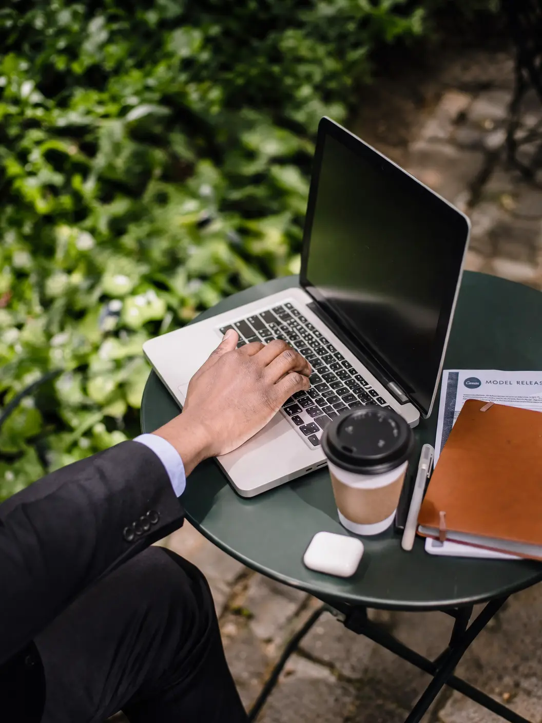 Hand typing on a laptop at an outdoor garden table with a coffee cup, notebook, and wireless earbuds case