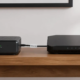 Black cable modem with four green status lights connected by an Ethernet cable to a flat black Wi-Fi router, both sitting on a wooden shelf beneath a framed picture on a white wall.