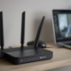 Black Wi-Fi 6 router with four antennas on a wooden desk next to an open laptop during a video call