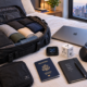 Open a black travel bag with neatly rolled clothes on a bed, beside a laptop, headphones, a phone, a portable Wi-Fi hotspot, a universal travel adapter, USA passport, and a journal reading "New places, Better me.